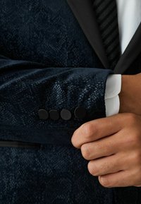 Jachetă bleumarin închis cu un model texturat, având patru nasturi negri la manșetă. Manșeta cămășii albe este vizibilă, suprapusă sub jachetă.