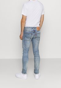 Hombre vistiendo jeans deslavados de color azul claro, zapatillas blancas y una camiseta blanca, de pie frente a un fondo blanco liso.