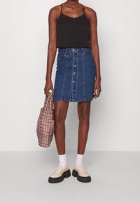 Kvinna i svart ärmlös topp, knäppt blå denimkjol, vita strumpor, beige plattform loafers, som håller en rutig väska i ena handen.