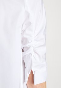 Camicia bianca con dettagli arricciati lungo le maniche, polsini a doppio bottone e una texture di tessuto liscia e croccante.