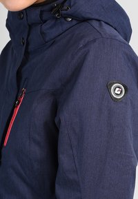Marineblaue Jacke mit Kapuze, ausgestattet mit einem roten Reißverschluss, strukturiertem Stoff und einem Logopatch auf der linken Schulter.