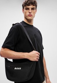 Μαύρη nylon τσάντα ώμου με μπροστινή τσέπη με φερμουάρ, λογότυπο "BOSS" σε λευκό, ρυθμιζόμενος ιμάντας και λεία υφή. Δομημένο σχήμα.