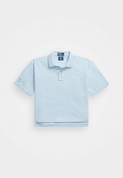 Šviesiai mėlynas polo marškinėlis, pagamintas iš medvilnės. Turi apykaklę, dvi mygtukų eiles ir mažą logotipą ant krūtinės. Trumpomis rankovėmis, viena iš jų su aplikacija.