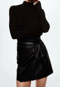 Personne portant un haut à col roulé noir à manches longues et une jupe portefeuille en similicuir noir avec un nœud noué sur le côté.