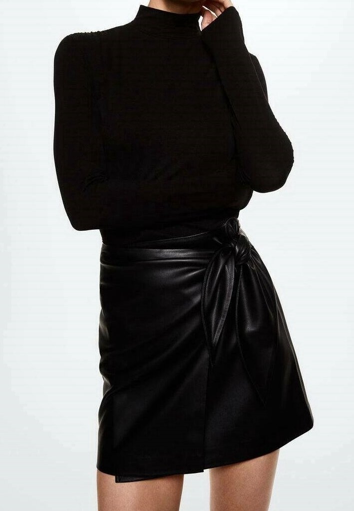 Personne portant un haut à col roulé noir à manches longues et une jupe portefeuille en similicuir noir avec un nœud noué sur le côté.