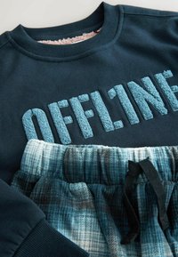 Pulover bleu cu glugă, având literele „OFFLINE” ridicate și pufoase; asortat cu pantaloni scurți vârsta cu carouri albastre și gri, dotati cu talie elastică și șnur negru.