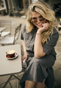 Femme blonde portant des lunettes de soleil et une robe grise, assise à une table de café en terrasse avec un latte, souriante et posant le menton sur la main.