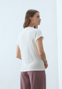 Lett hvit strikket topp med korte ermer, teksturert overflate og en subtil sømdetalj bak, kombinert med dempede mauve bukser.