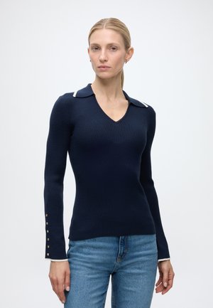 Kvinde iført en tætsiddende navy ribstrikket langærmet sweater med krave og guldknapper på manchetterne, kombineret med blå jeans mod en ensfarvet baggrund.
