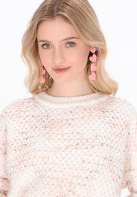 Fuchsia Herzohrringe in Tropfenform mit glänzendem Finish, bestehend aus drei geschichteten Herzen an goldfarbenen Ketten, getragen mit einem leicht strukturierten Pullover.