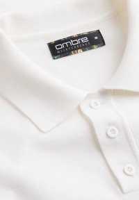 Gros plan sur un polo blanc avec un col plié, trois boutons blancs et une étiquette en tissu portant l’inscription "ombre QUINTESSENCE."