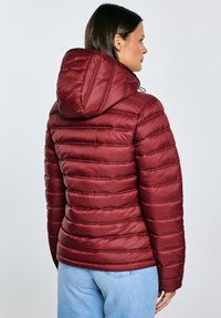 Žena v bordové prošívané bundě s kapucí, viděná zezadu, v kombinaci se světle modrými džínami na jednobarevném pozadí.