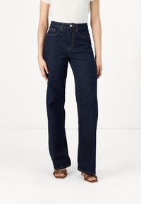 Femme portant un jean bleu foncé taille haute à jambes larges, un haut blanc à manches courtes et des sandales marron à talons.