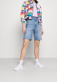 Kolorowa kurtka bomberka z mieszanym wzorem graficznym, zestawiona z jasnoniebieskimi szortami jeansowymi i białymi tenisówkami. Gładka tkanina i swobodny fason.