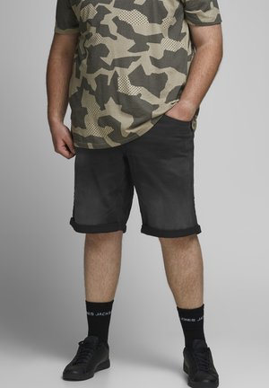 Hombre que lleva pantalones cortos negros remangados, una camiseta de camuflaje verde y negra, calcetines negros con texto blanco y zapatillas negras, de pie con las manos en los bolsillos.