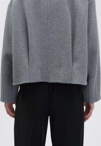 Grauer Wollpullover mit boxy Form, gesenkten Schultern und einem cropped Saum. Kombiniert mit glatten schwarzen Hosen. Weiche Textur ist deutlich zu erkennen.