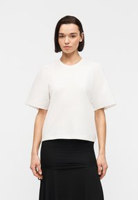 Γυναίκα με κοντά σκούρα μαλλιά, φορώντας λευκό μπλουζάκι με φουσκωτά μανίκια και μαύρη φούστα, στέκεται μπροστά σε απλό ανοιχτό γκρι φόντο.