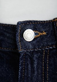 Mørkeblå denimstoff med brune sømmer. Har en sølvmetallknapp og en etikett med teksten 'gina uk'. Nærbilde av midjebåndet.