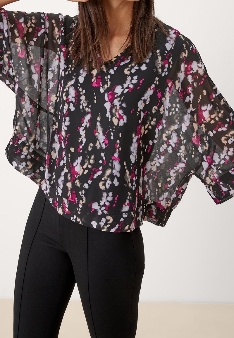 Femme portant un chemisier noir ample avec des motifs floraux abstraits roses, blancs et beiges, associé à un pantalon noir ajusté.