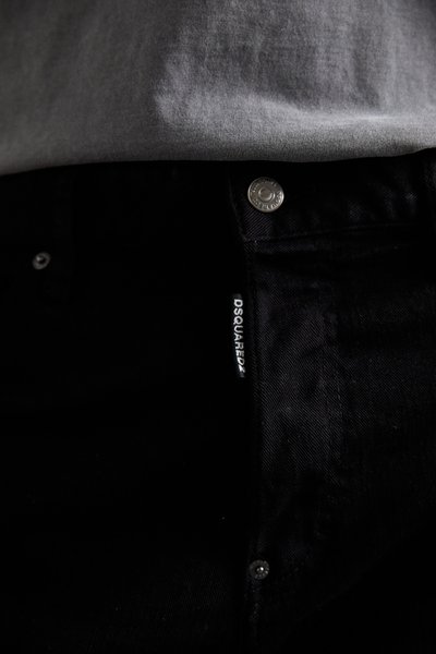 Nahaufnahme von schwarzen Jeans mit einem Metallknopf und dem sichtbaren DSQUARED2-Stoffetikett unter dem Saum eines grauen Shirts.