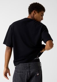 Czarny T-shirt z krótkim rękawem o luźnym kroju, o fakturze prążkowanej, noszony z ciemnymi dżinsami, z widocznym logo na pasie.