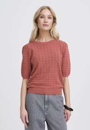 Femme blonde portant un pull court à manches courtes texturé de couleur rouille orangée et un jean taille haute gris clair, posant devant un fond uni.