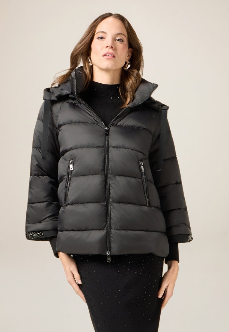 Czarna kurtka puchowa z zamkiem z przodu, dużym kapturem i szerokimi rękawami. Charakteryzuje się pikowaną fakturą i kieszeniami na zamek, skontrastowana z dopasowaną czarną sukienką.