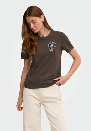Ung kvinde med langt hår iført brun t-shirt med sløjfegrafik og lyse beige bukser, der står mod en ensfarvet baggrund.