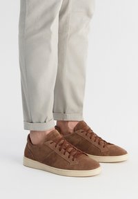 Brązowe zamszowe trampki z ciemnobrązowymi sznurówkami, o niskim profilu, zaokrąglonym czubku i minimalistycznym designie. Noszone z jasnymi, podwiniętymi spodniami.