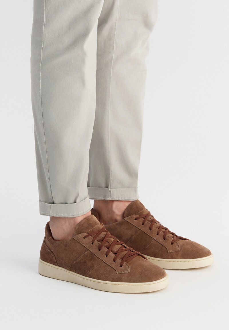 Brązowe zamszowe trampki z ciemnobrązowymi sznurówkami, o niskim profilu, zaokrąglonym czubku i minimalistycznym designie. Noszone z jasnymi, podwiniętymi spodniami.