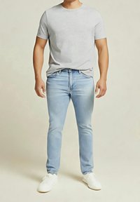Homme portant un t-shirt à manches courtes gris clair, un jean bleu clair et des baskets blanches, debout devant un fond uni.