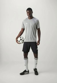 Camisa atlética de manga corta gris, pantalones cortos negros, calcetines blancos con rayas negras, sosteniendo un balón de fútbol con un diseño. Logo de Adidas en la ropa.