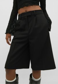 Pantalones cortos negros de corte ajustado, con un ajuste estructurado, que presentan pliegues, bolsillos laterales y una textura suave. Combinados con botas hasta la rodilla y calcetines blancos.