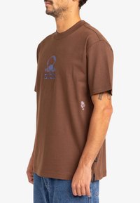 Brown cotton t-shirt featuring a blue logo design on the front and a small floral graphic on the sleeve, with short sleeves and a relaxed fit.