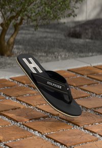 Tongs noires avec une semelle texturée, arborant un logo "H" blanc prominent et des coutures. Posées sur un motif de carreaux en bois et de gravier.