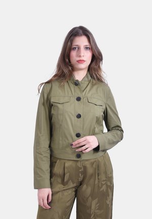 Giovane donna che indossa una giacca color verde oliva con bottoni e pantaloni coordinati a motivo floreale, in piedi davanti a uno sfondo chiaro e uniforme.