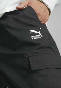 Pantalones cargo negros con una textura tejida, que presentan un logo de Puma en blanco y un bolsillo lateral. El diseño incluye un corte relajado para mayor comodidad.