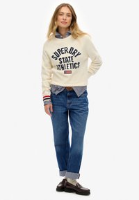 Pull crème avec impression navy "SUPERDRY STATE ATHLETICS," associé à une chemise col rayée et un jean bleu. Mocassins foncés et chaussettes beiges.