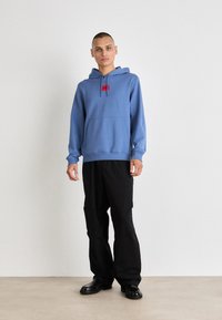 Felpa blu con un patch del logo rosso, tasca a canguro e cappuccio con cordino, abbinata a pantaloni neri a gamba larga e scarpe nere.