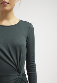 T-shirt à manches longues en tissu vert foncé avec un col rond et un détail torsadé à la taille, présentant une texture lisse et des manches ajustées.