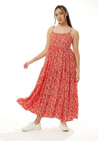 Red floral maxi dress with thin straps, gathered waist, and a flowing skirt. Worn with white sneakers; features small white daisies on fabric.