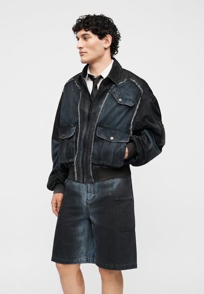 Νέος άνδρας με σγουρά μαλλιά φοράει μαύρο και σκούρο μπλε τζιν μπουφάν με πολλές τσέπες και αντίστοιχα μακριά τζιν σορτς.