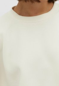 Crèmekleurige sweatshirt met een ronde hals, een gladde textuur en minimale naden, gepositioneerd tegen een effen achtergrond.