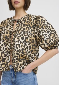 Blúzka s leopardím vzorom a puffovými rukávmi, s nastaviteľným viazaním vpredu a jemnou, štruktúrovanou tkaninou v zlatých a čiernych odtieňoch. Kombinovaná s džínsami.