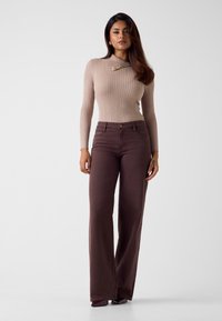 Maglione beige a coste con maniche lunghe, caratterizzato da un colletto decorativo e dettagli dorati. Abbinato a pantaloni marroni in denim a gamba larga.