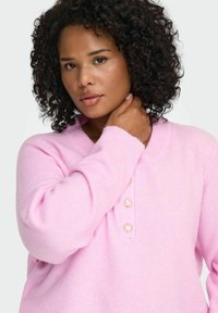 Rožinė megzta liemenė su apykakle, kuri turi širdelės formos mygtukus. Medžiaga atrodo minkšta ir tekstūruota, su šiek tiek per didelėmis rankovėmis.