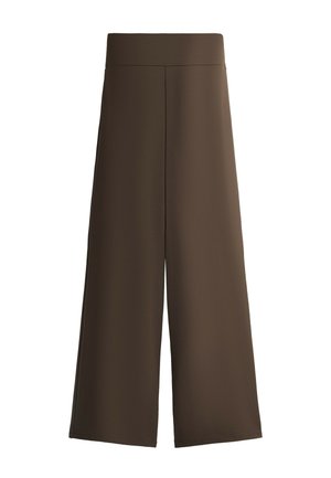 Frau trägt beige Kappe, marineblauen Mantel, hellgrauen Pullover, gestreiften Pullover um die Taille gebunden, braune Hose und Turnschuhe, steht vor grauem Hintergrund.