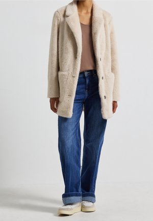 Henkilö, joka käyttää beigeä pörröistä takkia, ruskeaa toppia, sinisiä käännettyjä farkkuja ja valkoisia lenkkareita, seisoo yksinkertaisen vaalean taustan edessä.