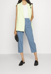 Nainen, jolla on vaaleanvihreä hihaton bleiseri, valkoinen toppi, siniset lyhyet housut ja mustat slip-on-kengät, pitää kädessään mustaa clutch-laukkua.