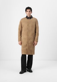Manteau en daim tan avec un col en cuir noir, manches longues, et deux poches latérales, porté sur un pantalon et des chaussures noirs.
